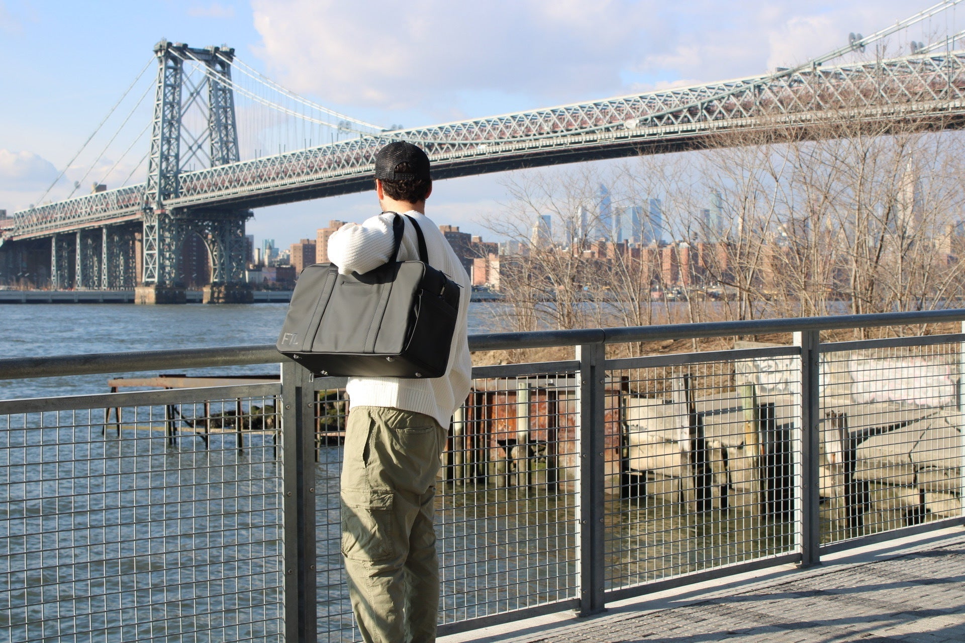 Gym duffle bag carried by man overlooking river with bridge and city skyline in the background.