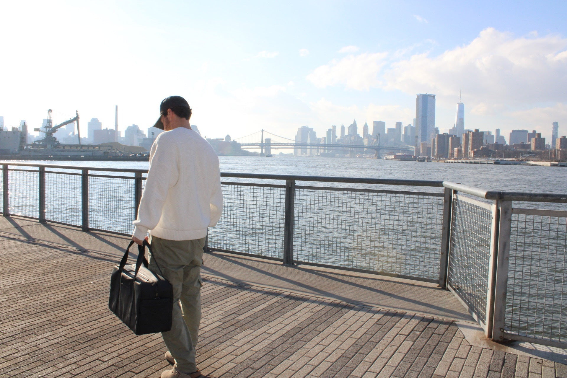Gym and work bag carried by a man walking along urban waterfront with the city skyline in the background.