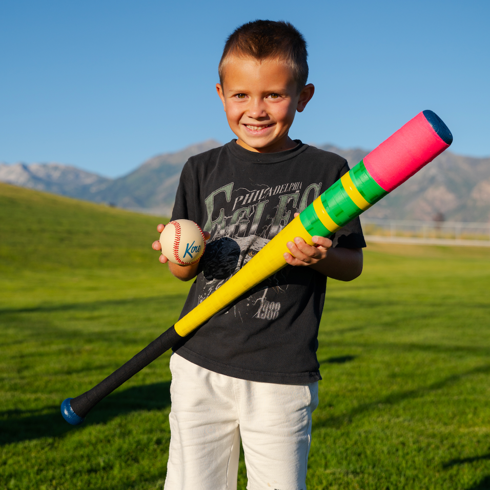 Child holding a pencil plastic wiffle baseball bat and Kore baseball on a grassy field with mountains in the background