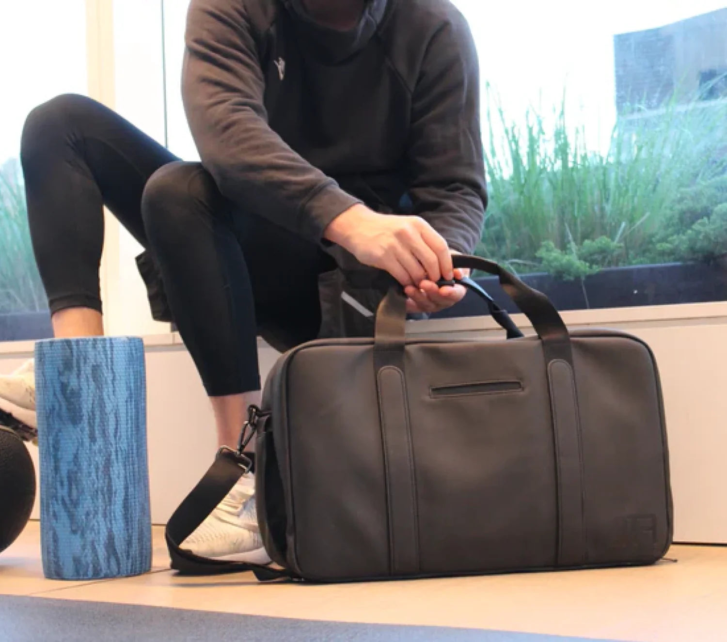 Men's gym duffel placed on floor next to foam roller and workout gear, handled by man in athletic wear.